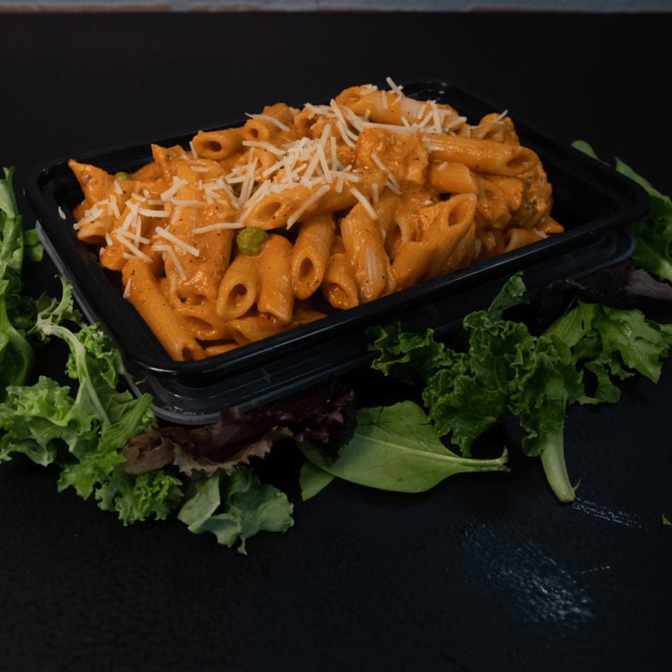 Penne pasta with creamy red sauce, shredded cheese, and peas in a black container with lettuce.